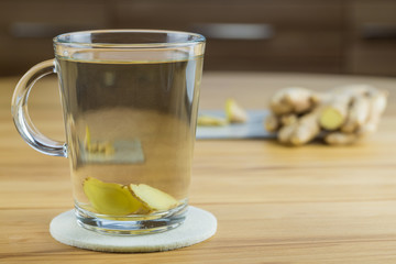 Ein Glas frischen Ingwerwassers, im Hintergrund sieht man eine Ingwerwurzel und in Scheiben geschnittenen Ingwer, das Schneidebrett mit den Ingwerscheiben spiegelt sich im Glas