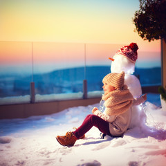 happy little kid and his friend snowman watching the sun goes down sitting in snow on rooftop...