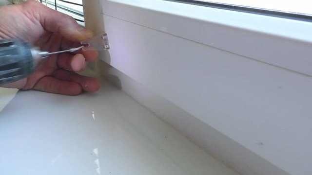 A man drills a hole in the window frame for fixing plastic bracket.
