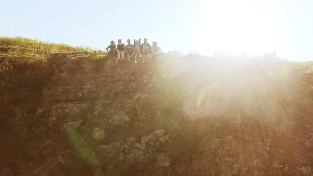 Group of happy tourists sitting on the cliff near precipice than waving hand and fall back on. View with tilt from drone in slowmotion