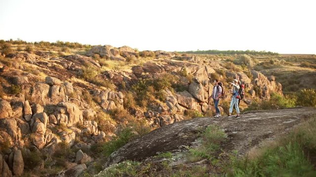Three friends walking in rocky landscape with camping backpacks giving high five each other. Panoramic view in slowmotion