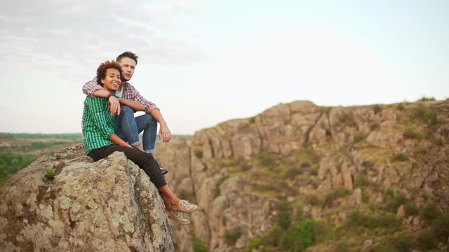 Caucasian male sitting with his African American girlfriend on rock admiring view smiling. He hugs her shoulders. In slowmotion