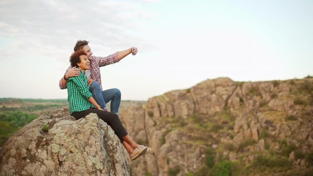 Interracial couple sitting on big rock making selfie with mobile phone smiling posing in slowmotion