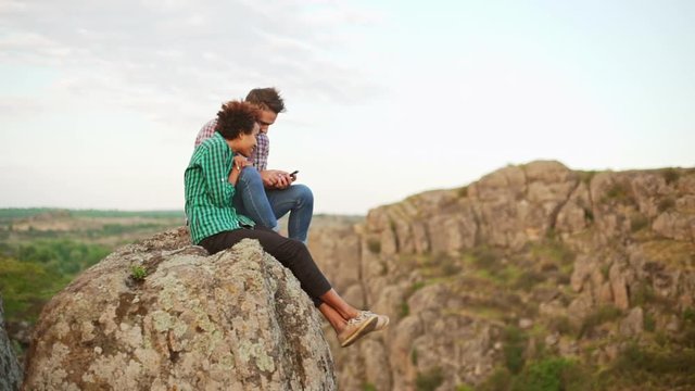Beautiful interracial pair sitting on cliff looking photos on mobile phone laughing discussing swiping in slowmotion