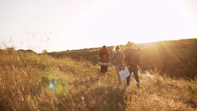 Three tourists walking away with camping backpacks and map through yellow prairie. Back view in slowmotion