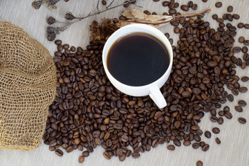 White cup of coffee standing on coffee beans, poured out of the bag on a gray tablecloth