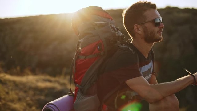 Caucasian bearded male in black sunglasses and backpack with sleeping pad sitting outdoors while sun shining on background in slowmotion