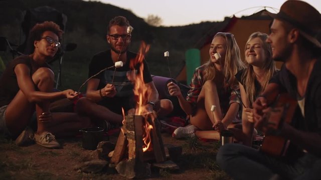 Friends sitting near bonfire near tent camps at night eating marshmallow playing guitrar smiling laughing talking in slowmotion