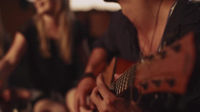 Happy Caucasian male with light bristle and hat playing guitar while two blonde female smiling on background. Close up in slowmotion