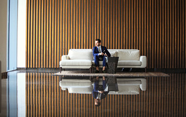 Happy young businessman sitting relaxed on sofa at hotel lobby using smartphon, waiting for someone.