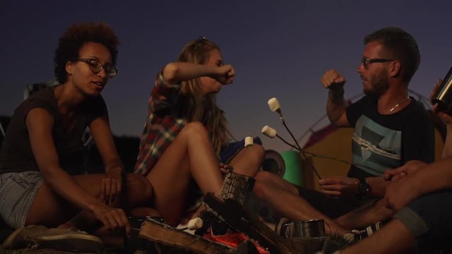 Blonde beautiful female in plaid shirt giving fist bump to bearder caucasian male in glasses outdoors near fire while dark-skinned girl siiting near them. In slowmotion