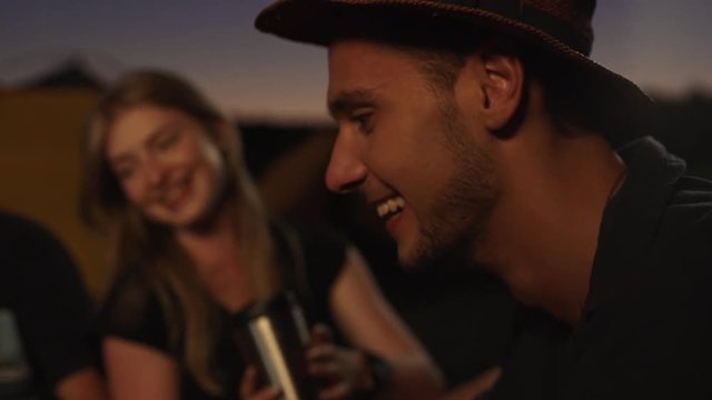 Handsome Caucasian male with light bristle sitting in hat smilling talking with blonde beautiful girl on background with thermo cup. Close up in slowmotion