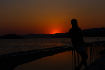 Fototapeta premium silhouette of a girl and basket flowers standing on the bank wide river in the rays the setting warm, beautiful sun