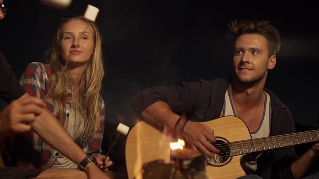 Young handsome Caucasian male playing guitar smiling while blonde beautiful female listening enjoying music. In slowmotion