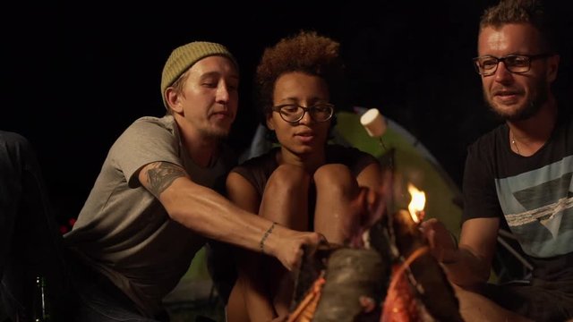 Dark-skinned girl in glasses sitting among two Caucasian male in glasses eating fried marshmallows near fire outdoors. In slowmotion