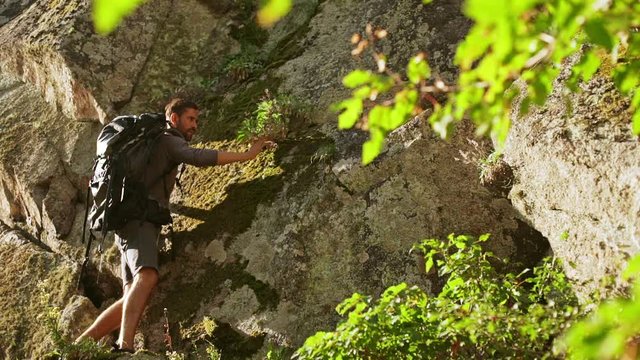 Brave Caucasian male with dark beard climbing up rock wearing sandales and tourist backpack. In slowmotion