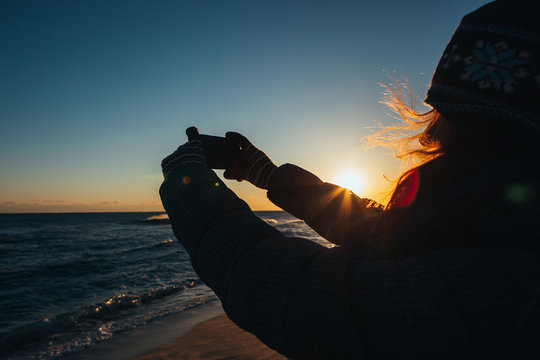 Girl makes a photo on your phone. Her hair fluttering in the wind, she is dressed in winter clothes. Sunrise in the ocean. The glare of the sun illuminate the silhouette.