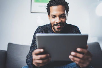 Selective focus.Attractive bearded African man using tablet while sitting on sofa in his modern office.Concept of young business people working at home .Blurred background.