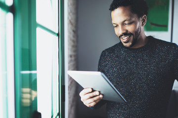 Selective focus.Smiling bearded African man reading book on digital tablet while standing near the window in his modern apartment.Concept of young business people working at home.Blurred background.
