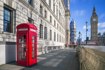 Fototapeta na wymiar London, England - Traditional red british telephone box with Big Ben and Double Decker bus at the background on a sunny afternoon with blue sky and clouds