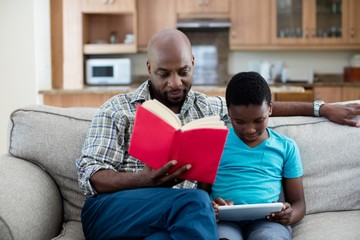 Father reading book while son sitting next to him using digital 