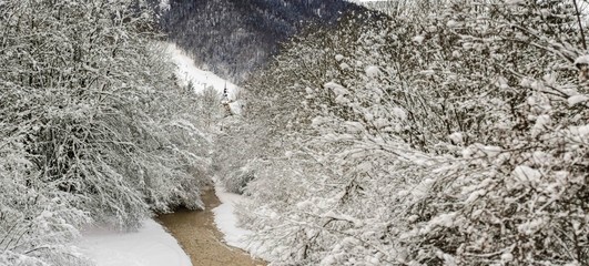 Panoramic view of Alpine river surrounded by snowy branches with church tower and mountain in background.