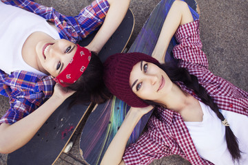 Two Brunette teenage girls friends in hipster outfit (jeans shorts, keds, plaid shirt, hat) with a skateboard at the park outdoors. © AnnaDemy