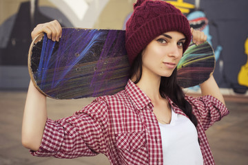 Brunette teenage girl in hipster outfit (jeans shorts, keds, plaid shirt, hat) with a skateboard at the park outdoors © AnnaDemy
