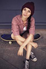 Brunette teenage girl in hipster outfit (jeans shorts, keds, plaid shirt, hat) with a skateboard at...