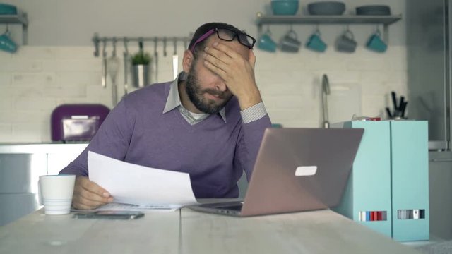 Overwhelmed, sad businessman with documents and laptop sitting by table in the kitchem, 4K
