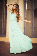 Beautiful blonde with a long curly hair in a long evening dress in motion outdoors near retro vintage building all in leaves in summer sunset