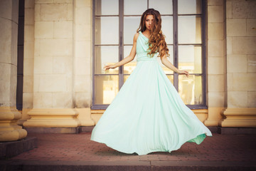 Beautiful blonde with a long curly hair in a long evening dress in motion outdoors near retro vintage building in summer sunset. Copy Space