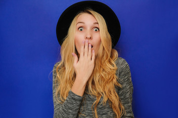 Caucasian blond girl in a black hat closes her mouth in surprise, shame, fear, horror. Blonde girl hipster in hat and gray sweatshirt on a blue background in shock from seasonal sales, big discounts.