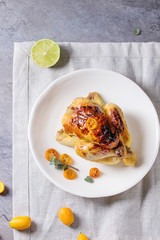 Whole roasted mini chicken cooking with caramelized kumquats served on white plate with textile napkin and fresh citrus fruits over gray kitchen table. Top view