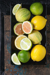 Variety of whole and sliced citrus fruits pink tiger lemon, lemons and lime, on black plate over dark ornate and old wooden background. Top view. Healthy eating