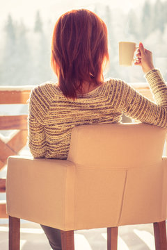 Woman Sitting In A Comfortable Chair Drinking Coffee And Looking Through Window At Snow Covered Mountain.