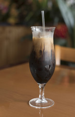 Tall shapely glass of Thai iced coffee with a straw on a brown wood table against a blurred restaurant background