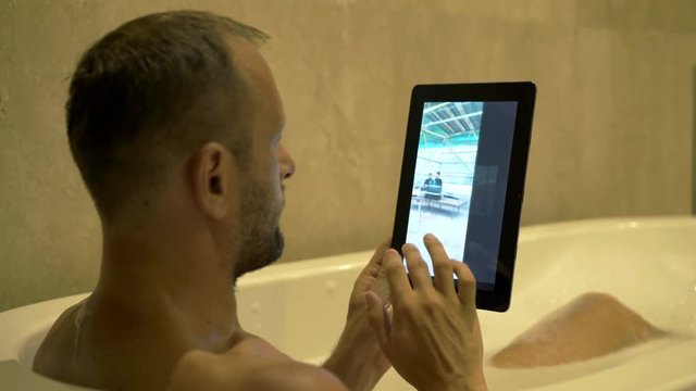 Young man browsing photos on tablet during bath, 4K
