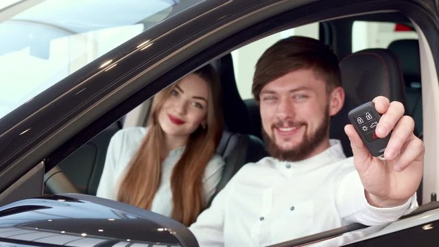 Close up of attractive caucasian man showing car key through the window. Nice young couple sitting inside the new car at the dealership. Handsome bearded man sticking his hand out of the vehicle