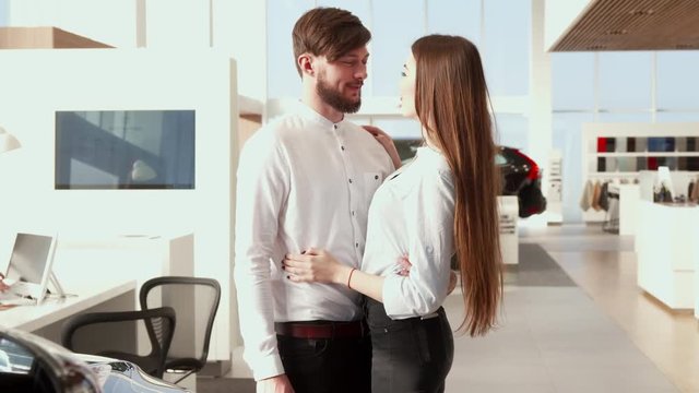 Brunette bearded man pointing his hand on the car at the dealership. Young caucasian man consulting with his pretty girlfriend at the car showroom. Attractive brown haired girl agreeing with her