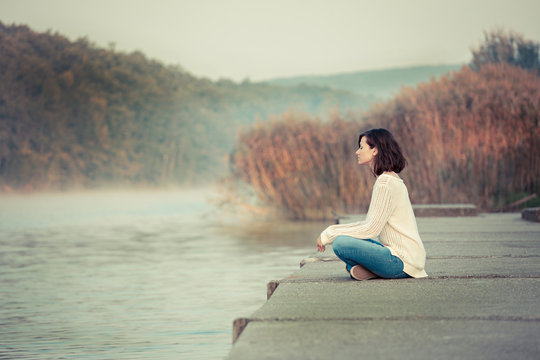 Relaxation by lake. Young woman sitting and looking away. Chilly morning. 