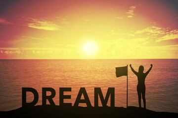 silhouette of winning success woman at sunset or sunrise standing and raising up hand near flag with text DREAM in celebration of having reached mountain top summit goal.business success concept