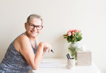 Older woman with short grey hair and glasses holding pen and smiling while sitting at writing desk with pink roses (selective focus)