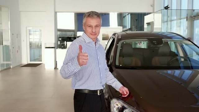 Attractive mature man showing his thumb up near the car at the dealership. Middle aged gray client approving new black SUV. Caucasian male client holding his hand on the car hood