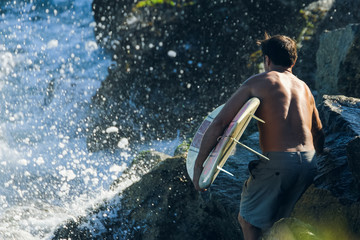 Man holding surfboard climbs over rocks as waves crash and spray ahead of him