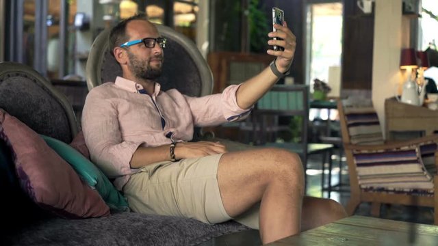 Young man taking selfie photo with cellphone sitting on sofa in trendy cafe
