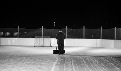 An adult male shovelling off an outdoor hockey rink at night under lights in black and white