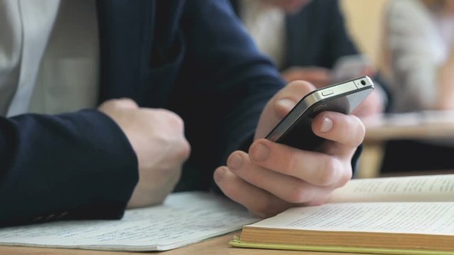 The student sitting at the school desk at school writes the text using the cellphone indoors