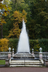 Pyramid Fountain at Peterhof with dirt path and stone steps leading to the fountain, stone banisters around the fountain and fall foliage in the background.