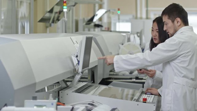 Tracking shot of young man and woman in lab coats standing at SMT machine and discussing something at computer screen; woman using digital tablet